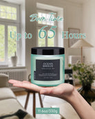 Candle with a label held by hands against a room background, featuring text about burn time and weight.