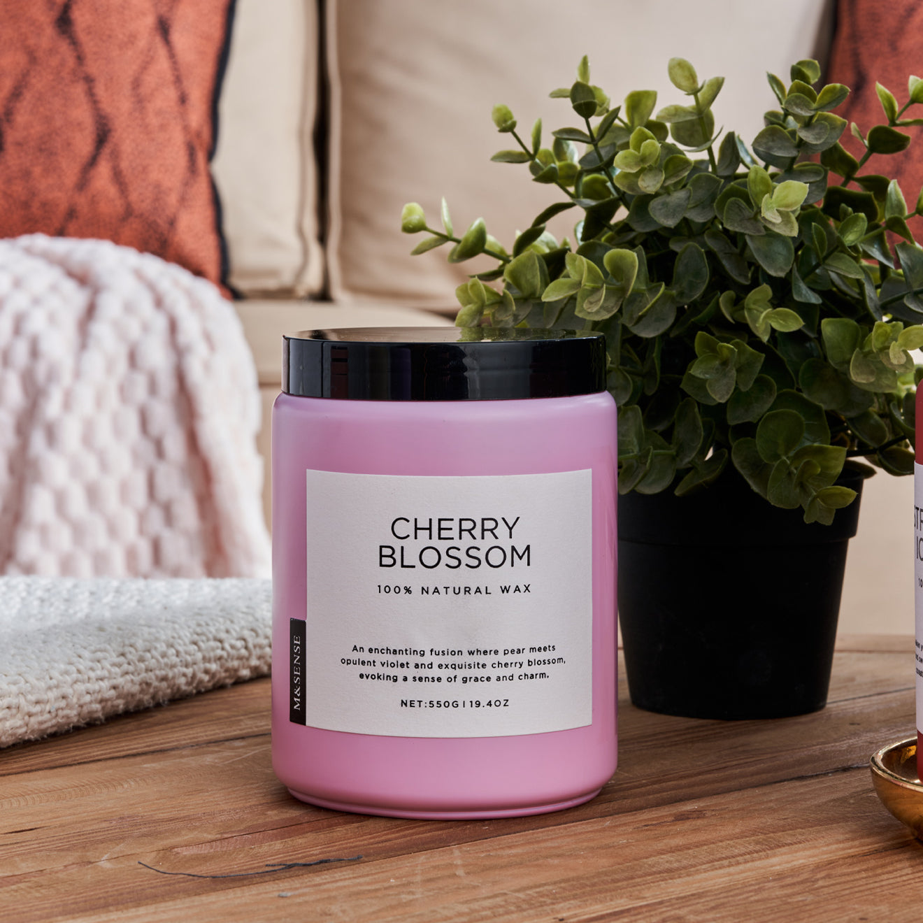 Cherry Blossom scented candle on a wooden table with a green plant, against a sofa background.