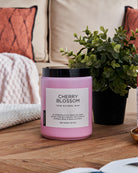 Cherry Blossom scented candle on a wooden table with a green plant, against a sofa background.