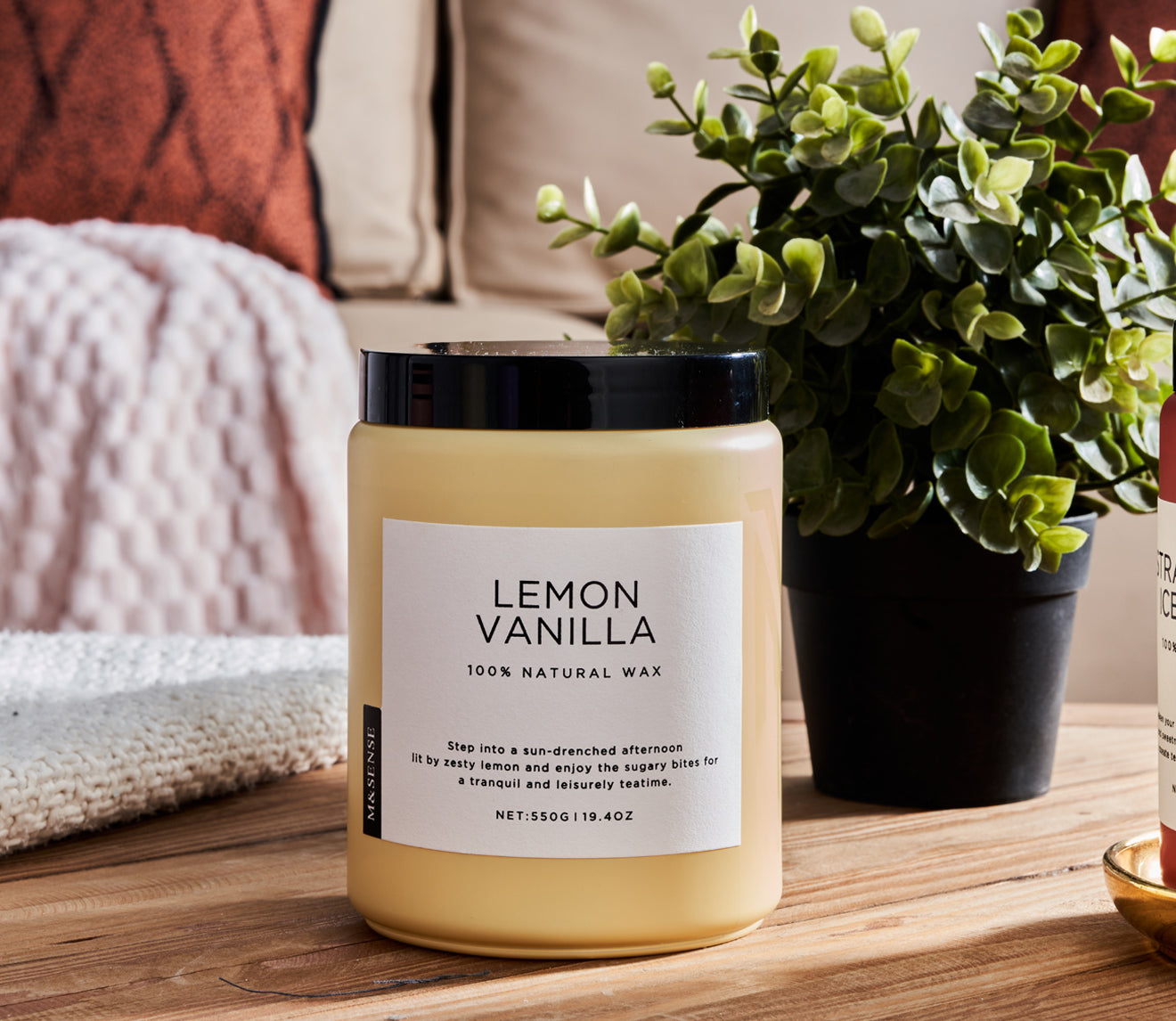 Lemon Vanilla scented candle on a wooden table with a green plant, against a sofa background.