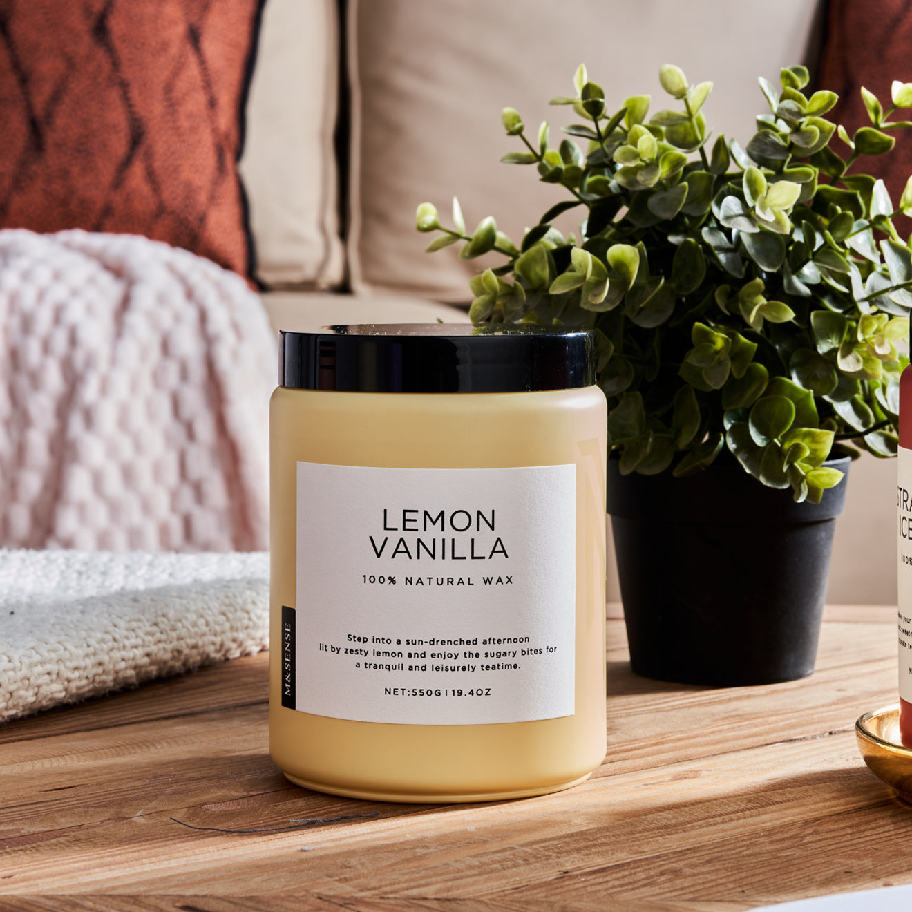 Lemon Vanilla scented candle on a wooden table with a green plant, against a sofa background.