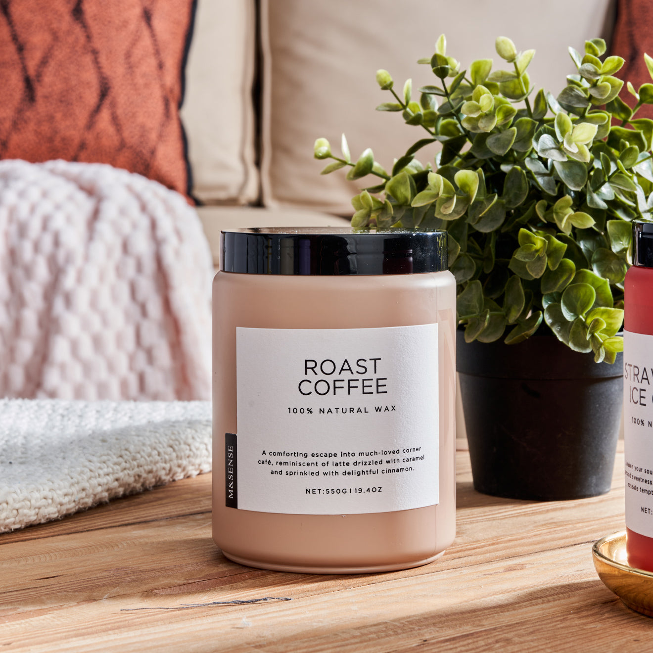 Pink Roast Coffee scented candle on a wooden table with a green plant, against a sofa background.