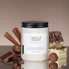 Vanilla cream candle with cinnamon sticks, chocolate, and a slice of cake on a reflective surface.
