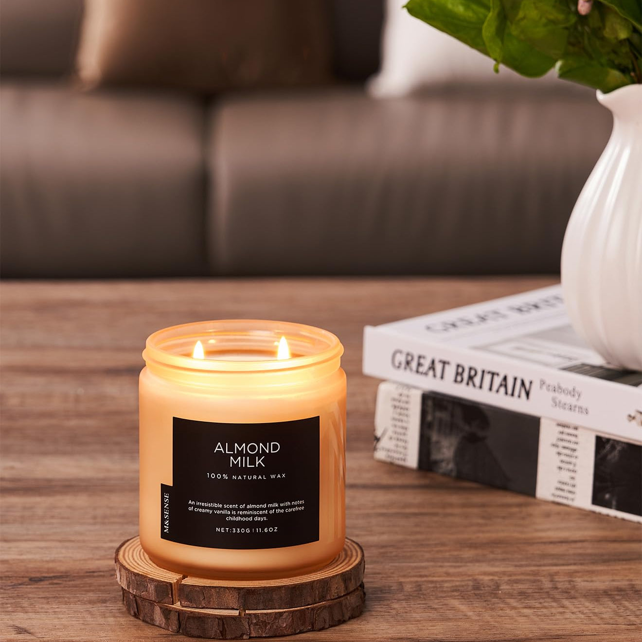 Candle labeled 'Almond Milk' on a wooden coaster with a blurred background of a couch and plant.