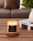 Candle labeled 'Almond Milk' on a wooden coaster with a blurred background of a couch and plant.