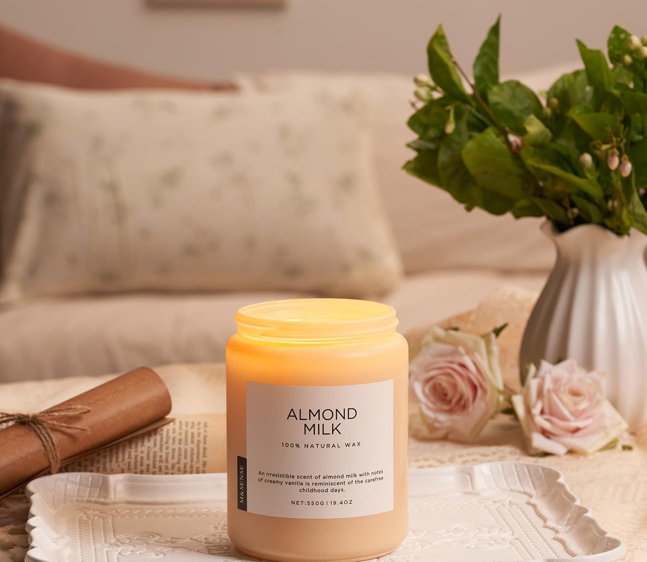 Candle labeled 'Almond Milk' on a table with flowers and a vase in the background