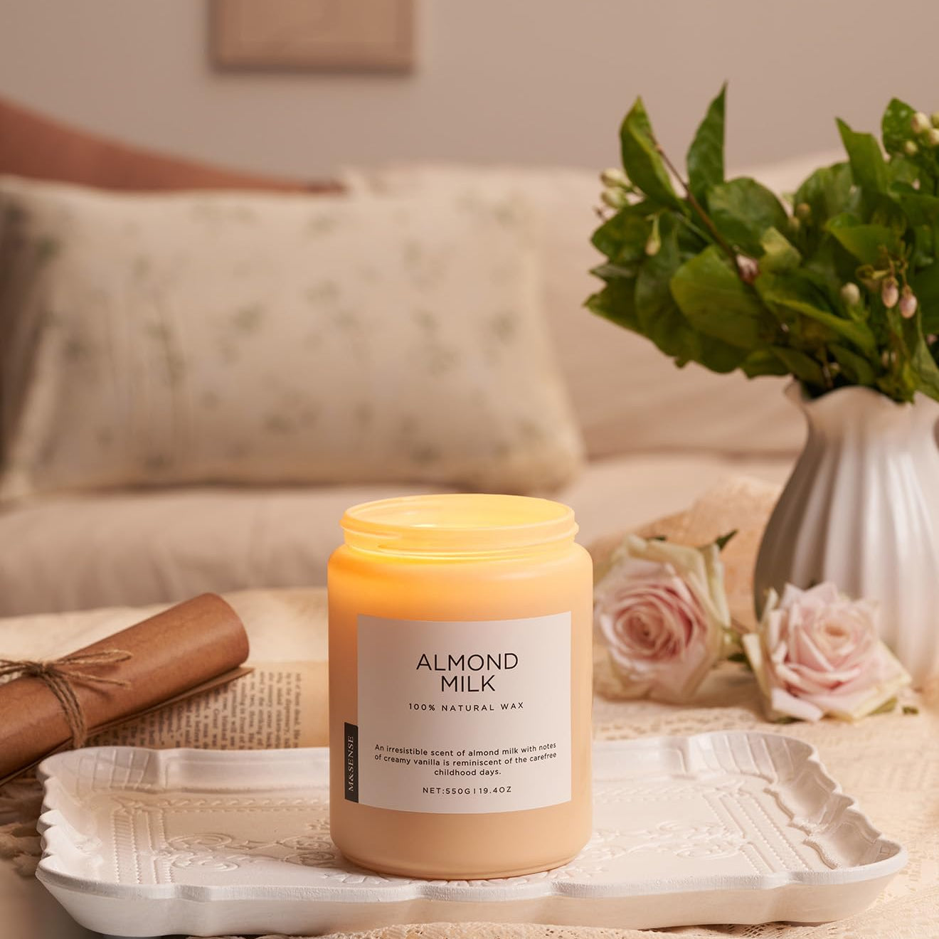 Candle labeled 'Almond Milk' on a table with flowers and a vase in the background