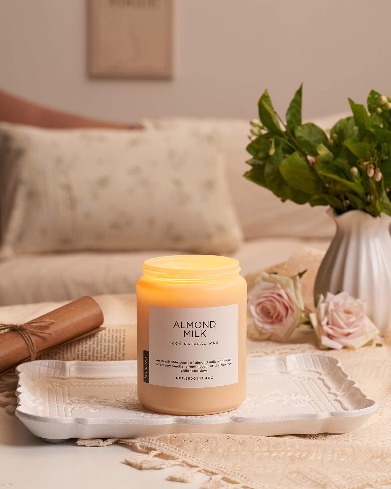 Candle labeled 'Almond Milk' on a table with flowers and a vase in the background
