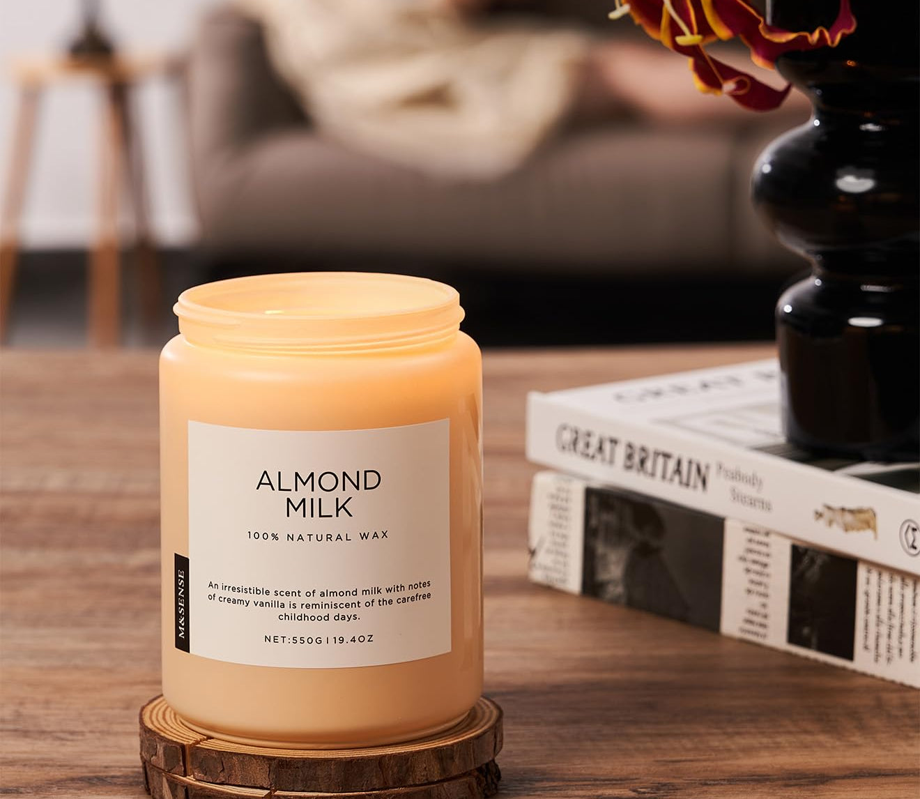 Candle labeled 'Almond Milk' on a wooden coaster with a blurred background of a living room.