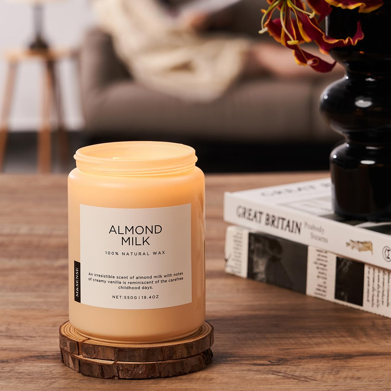 Candle labeled 'Almond Milk' on a wooden coaster with a blurred background of a living room.