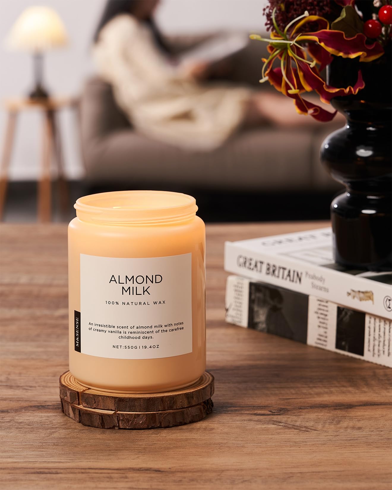 Candle labeled 'Almond Milk' on a wooden coaster with a blurred background of a living room.
