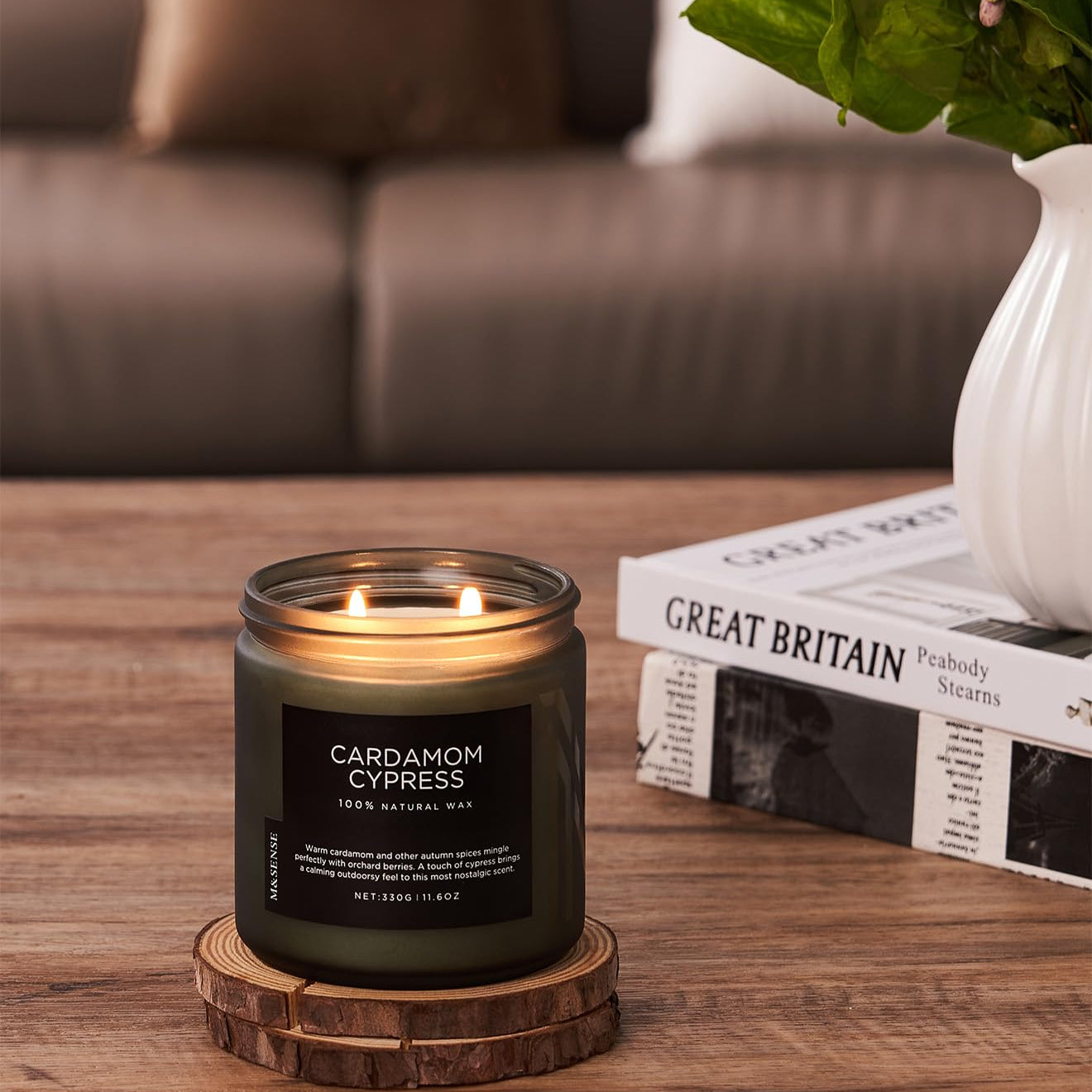 Candle labeled 'Cardamom Cypress' on a wooden coaster with a blurred sofa and plant in the background.