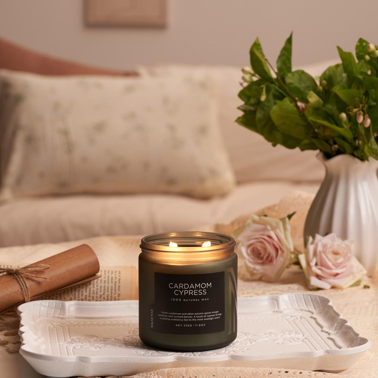 Candle labeled 'Cardamom Cypress' on a tray with flowers and a vase in the background