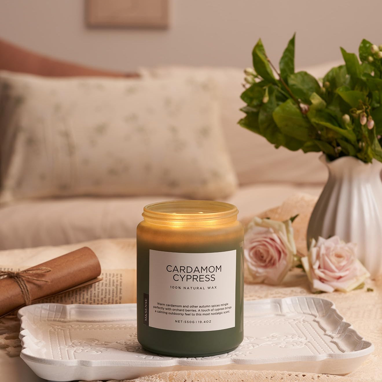 Candle labeled 'Cardamom Cypress' on a table with flowers and a vase in the background