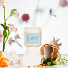 Candle labeled 'Coconut Lime & Apricot' with a coconut shell and fruits on a white surface with flowers in the background.