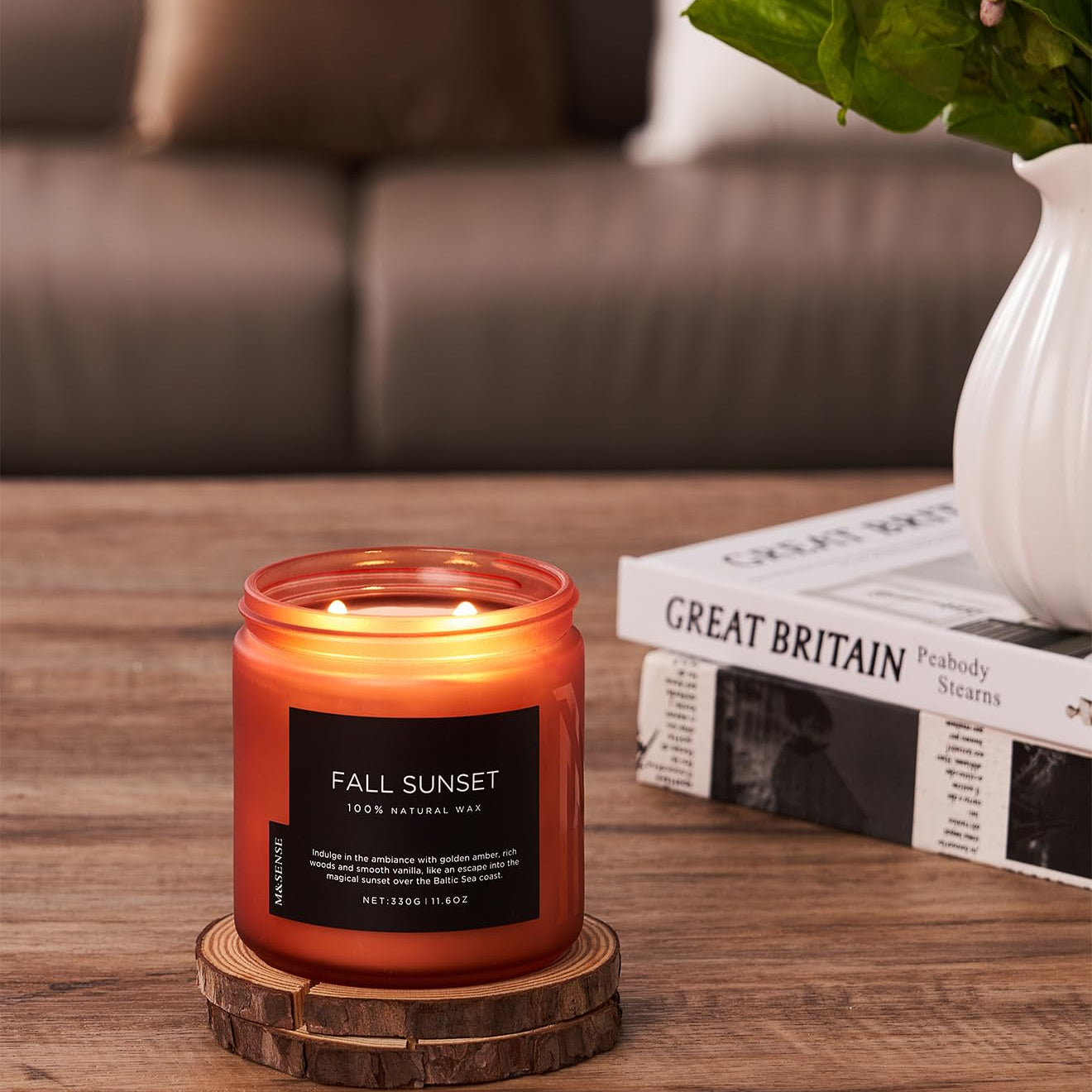 Candle labeled 'Fall Sunset' on a wooden coaster with a blurred background of a couch and books.