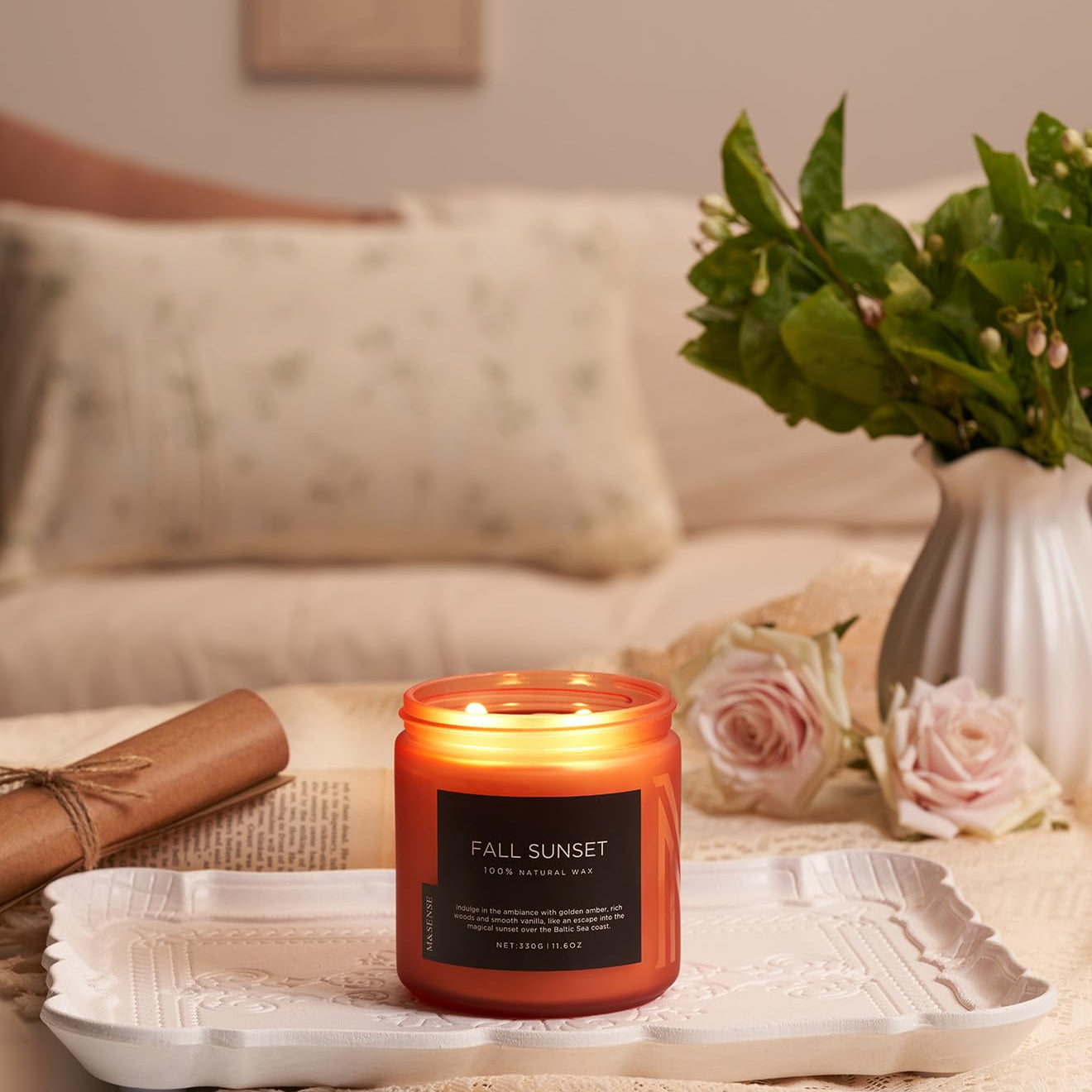 Candle labeled 'Fall Sunset' on a table with flowers and a vase in the background