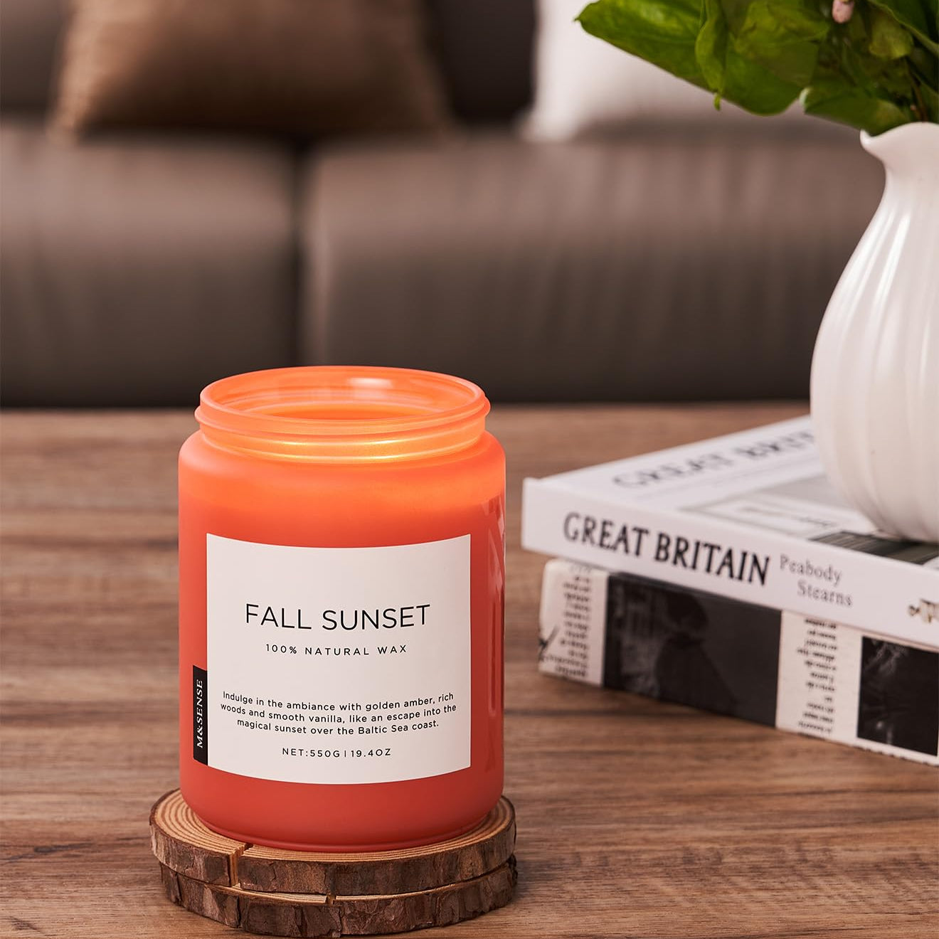 Orange candle labeled 'Fall Sunset' on a wooden coaster with a magazine and plant in the background.