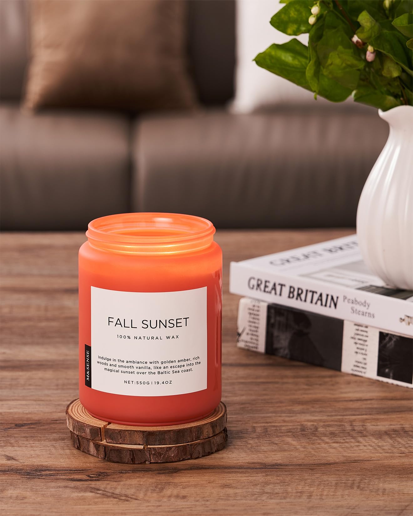 Orange candle labeled 'Fall Sunset' on a wooden coaster with a magazine and plant in the background.