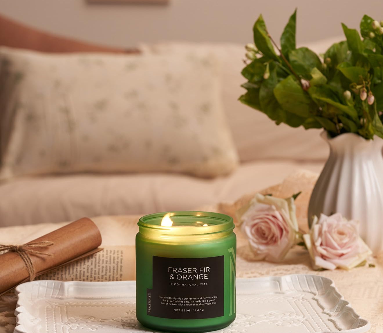 Green candle labeled 'Fraser Fir & Orange' on a tray with flowers and a vase in the background.