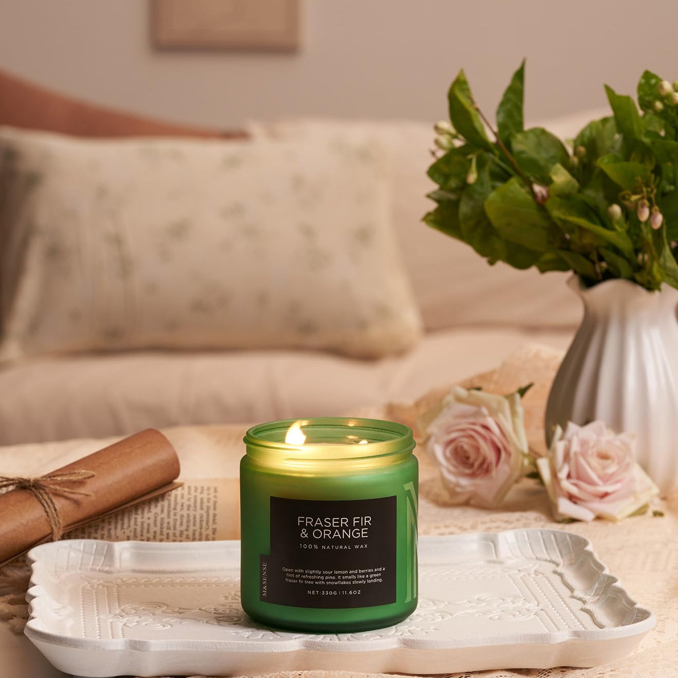 Green candle labeled 'Fraser Fir & Orange' on a tray with flowers and a vase in the background.