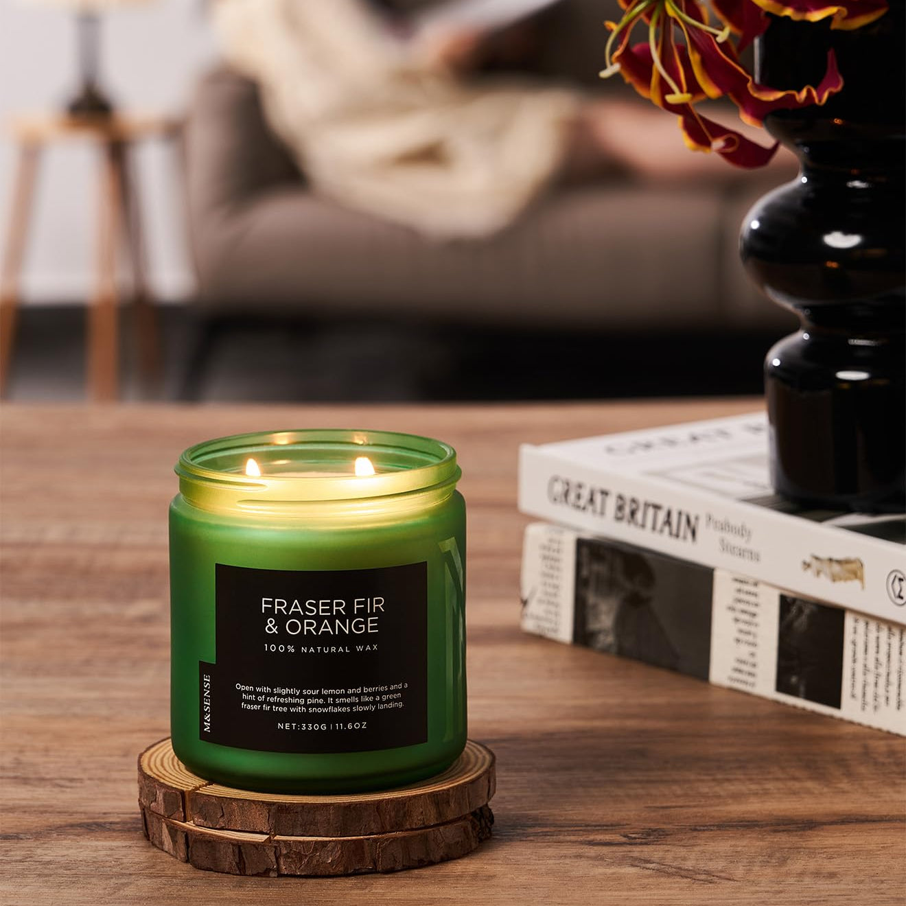 Green candle labeled 'Fraser Fir & Orange' on a wooden coaster with a blurred background of a cozy living room.