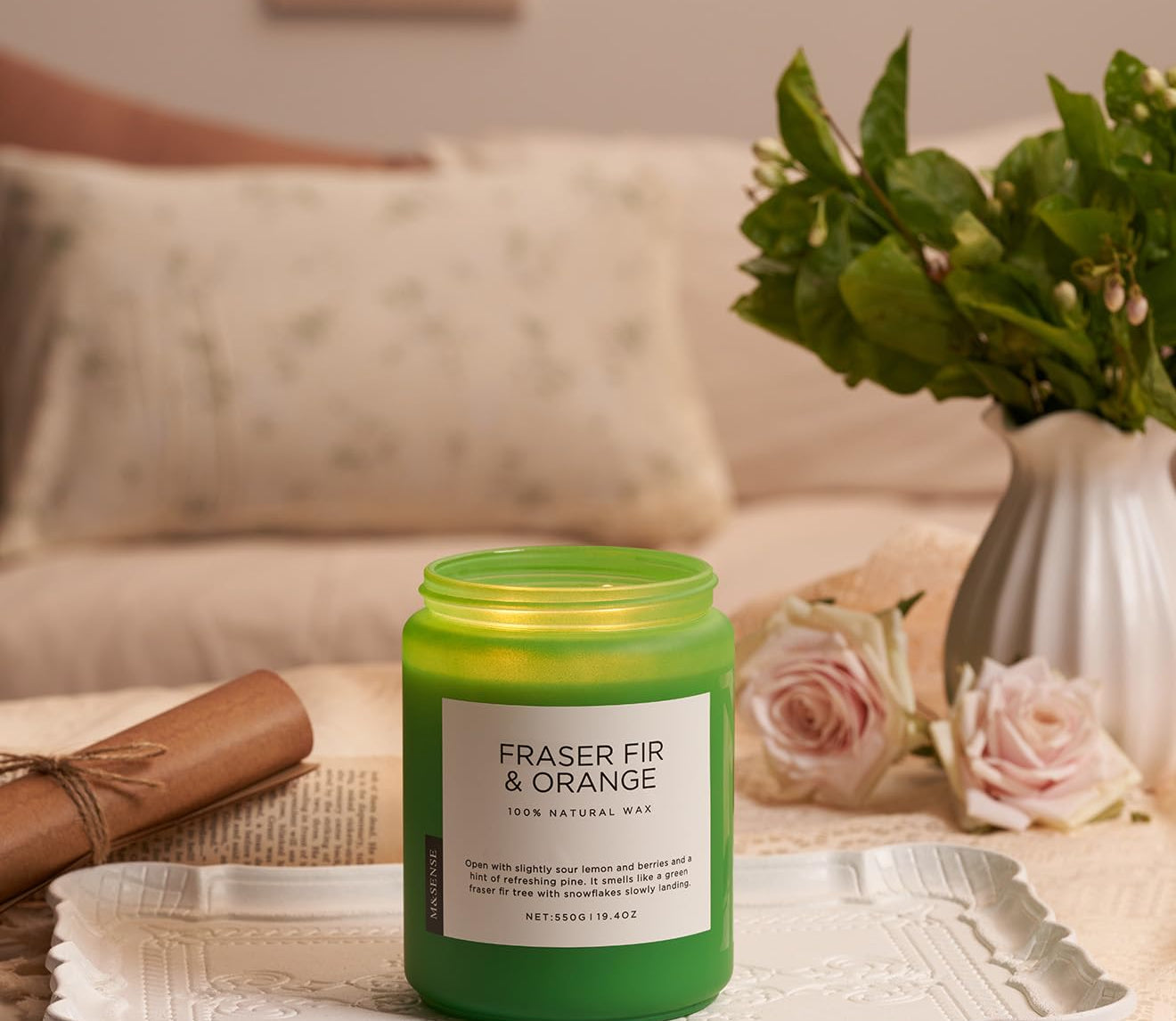 Green candle labeled 'Fraser Fir & Orange' on a tray with flowers and a vase in the background.