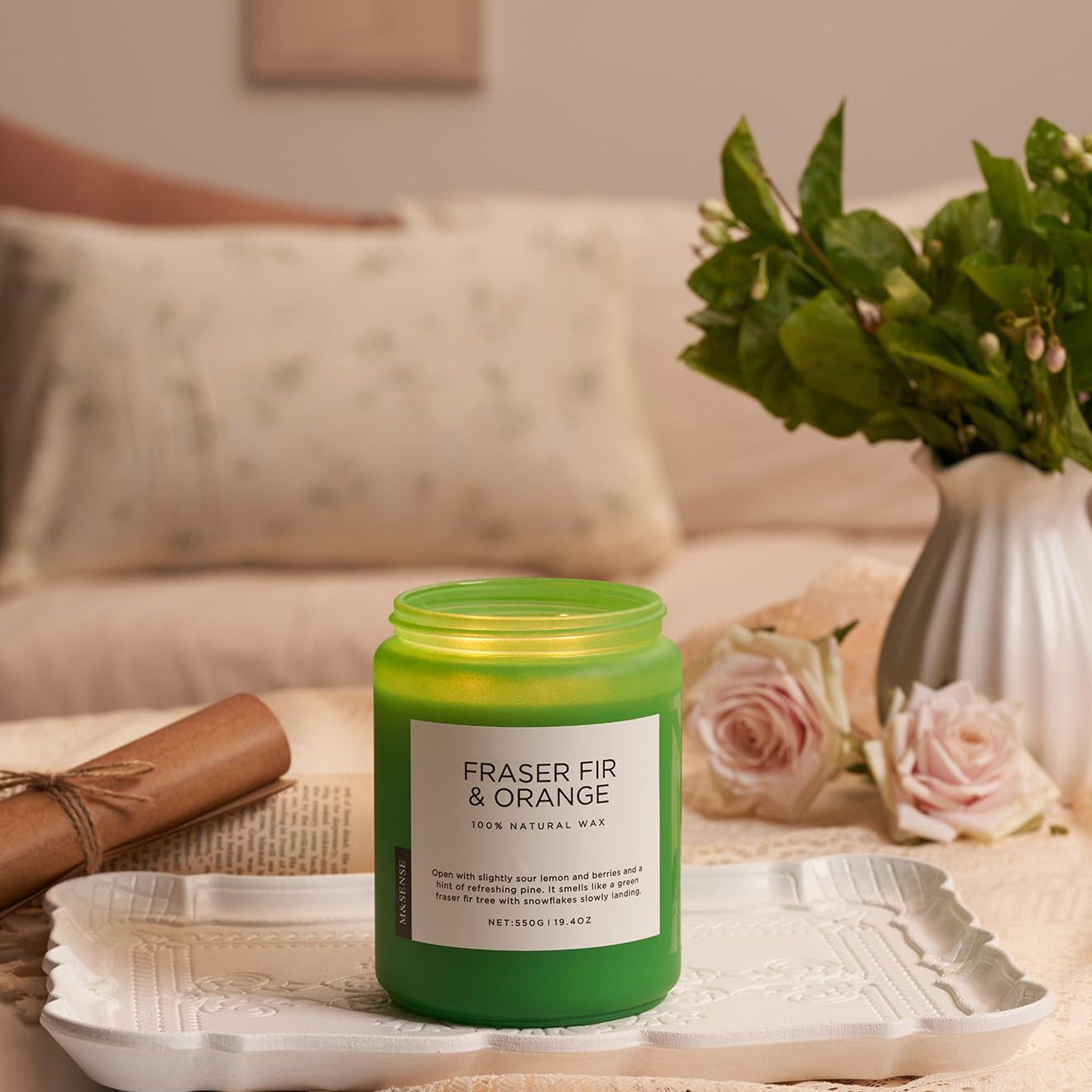 Green candle labeled 'Fraser Fir & Orange' on a tray with flowers and a vase in the background.