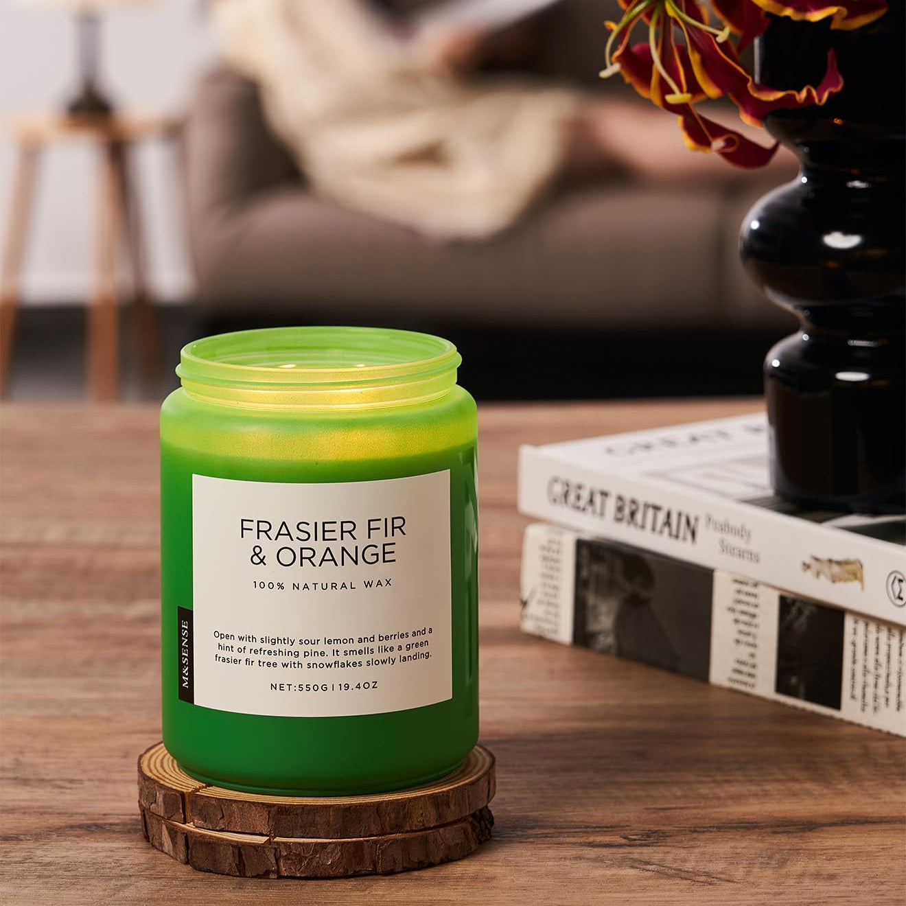 Green candle labeled 'Frasier Fir & Orange' on a wooden coaster with a blurred background of a person reading.