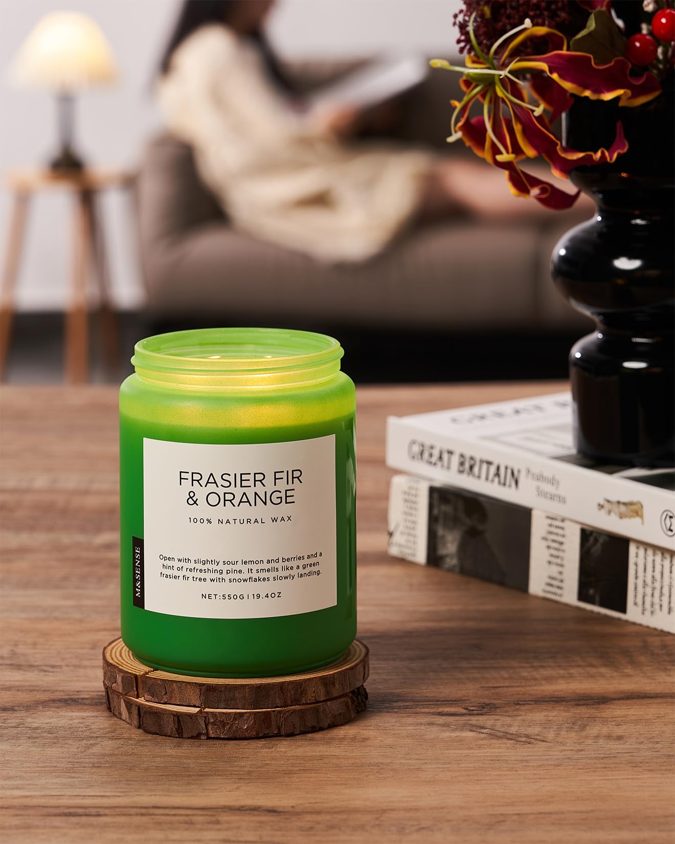 Green candle labeled 'Frasier Fir & Orange' on a wooden coaster with a blurred background of a person reading.