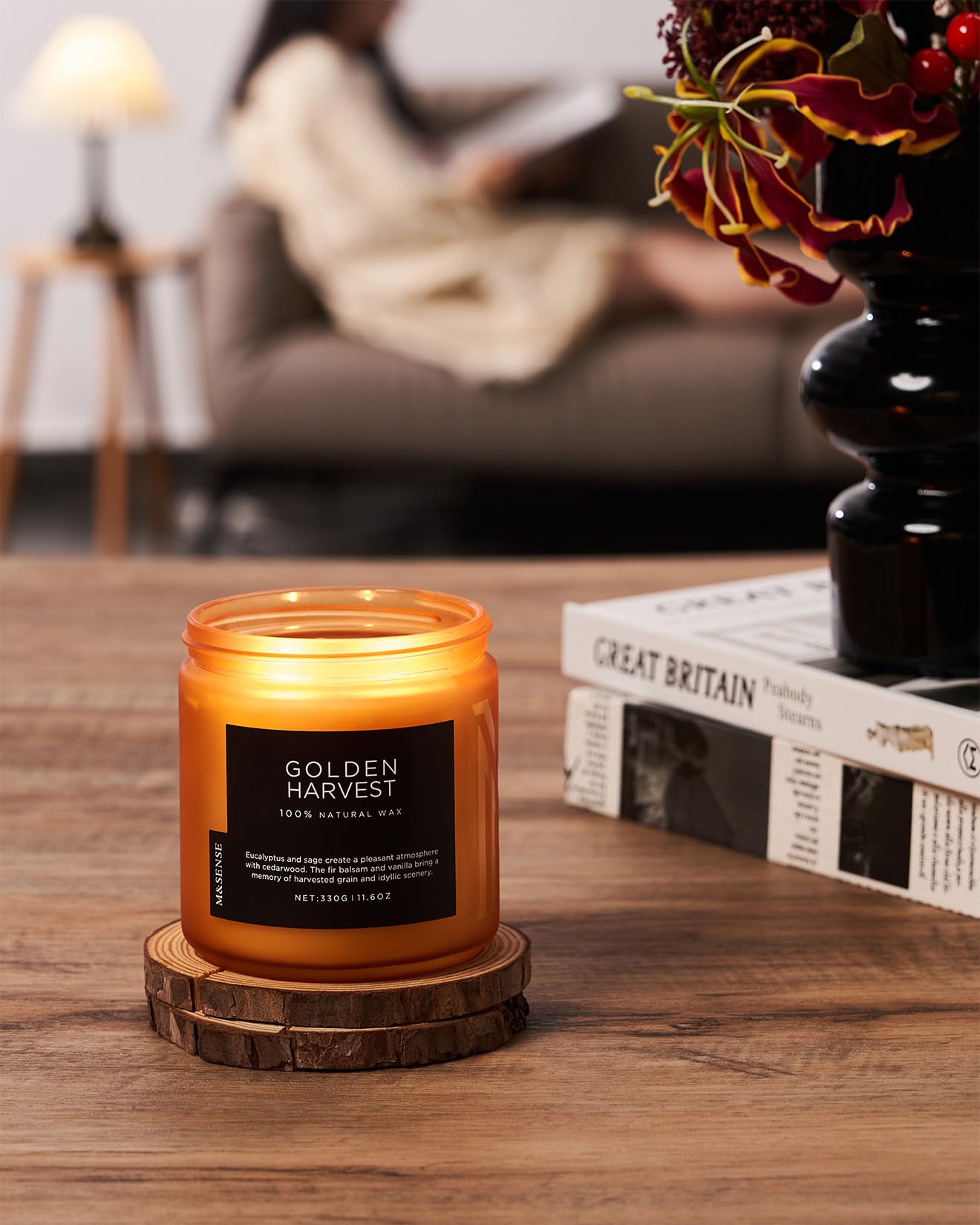 Candle labeled 'Golden Harvest' on a wooden coaster with a blurred background of a person reading.