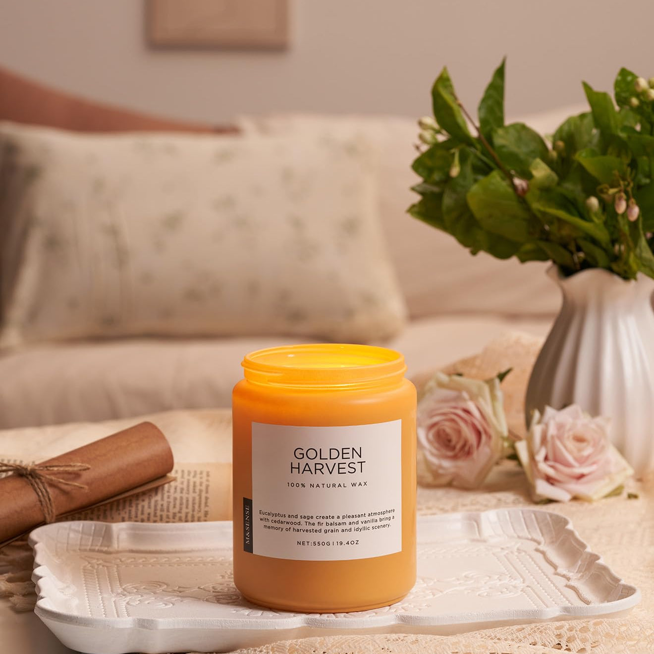 Candle labeled 'Golden Harvest' on a table with flowers and a couch in the background