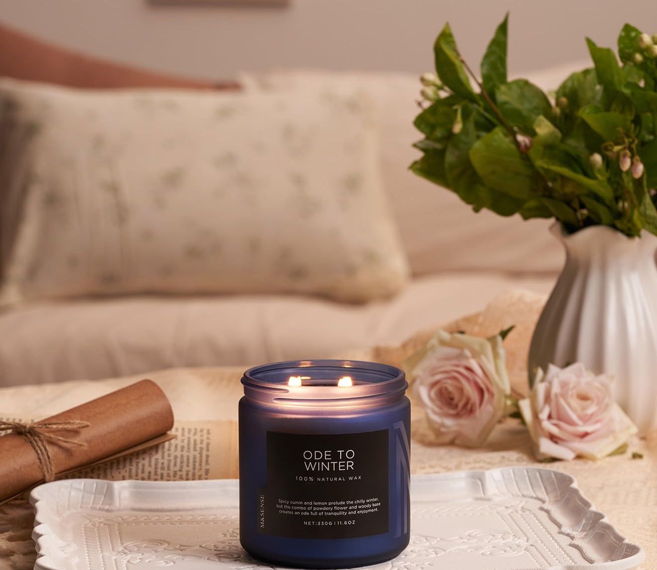 Candle labeled 'Ode to Winter' on a decorative tray with flowers and pillows in the background