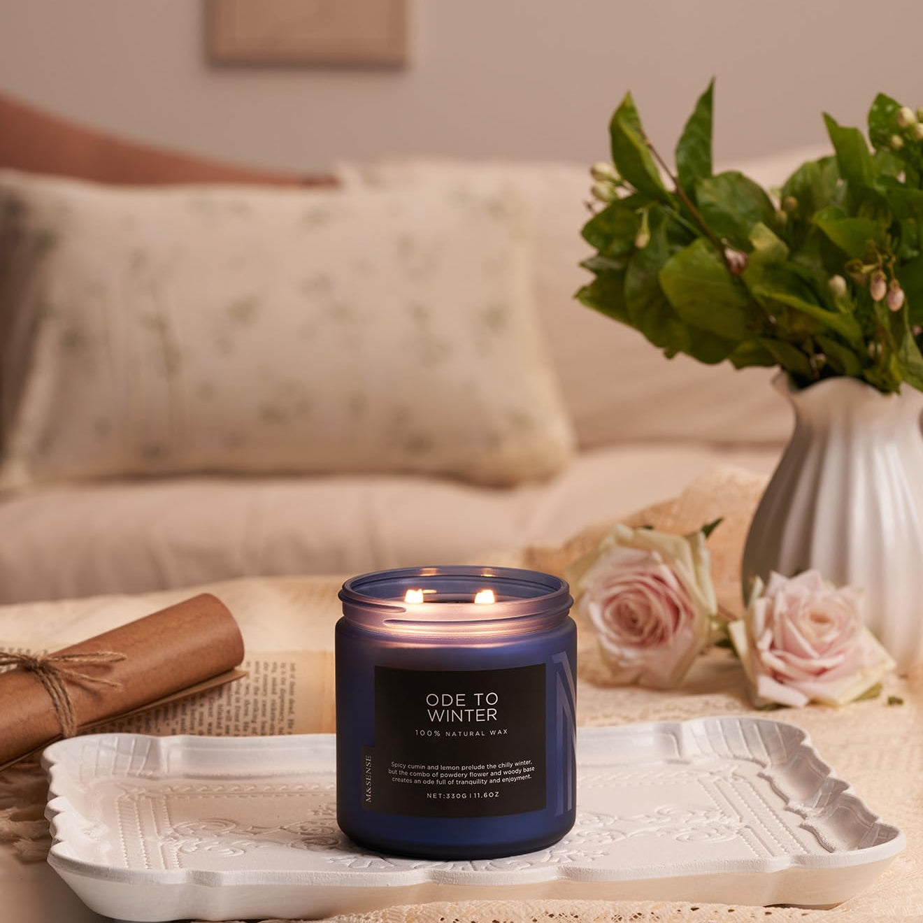 Candle labeled 'Ode to Winter' on a decorative tray with flowers and pillows in the background