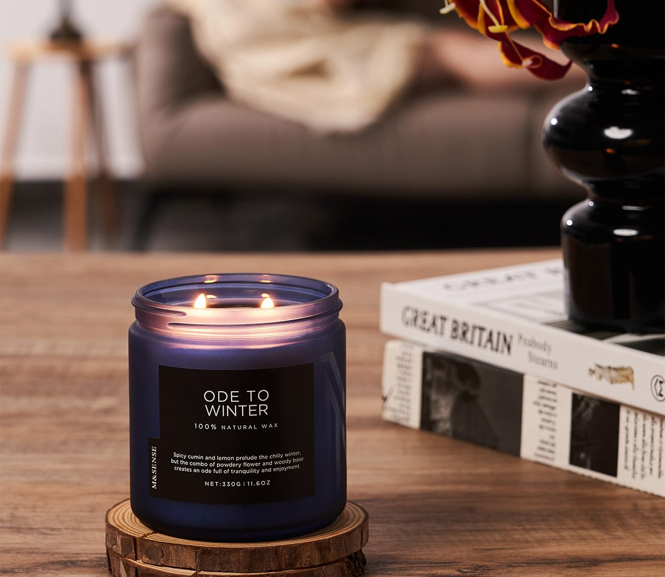 Candle labeled 'Ode to Winter' on a wooden coaster with a blurred background of a person reading.
