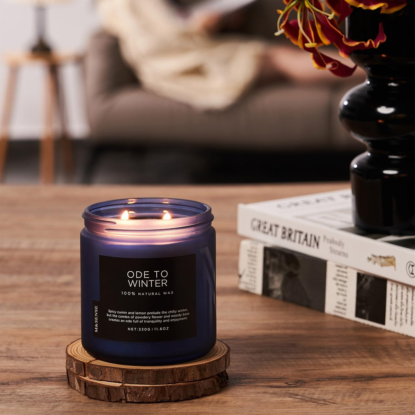 Candle labeled 'Ode to Winter' on a wooden coaster with a blurred background of a person reading.