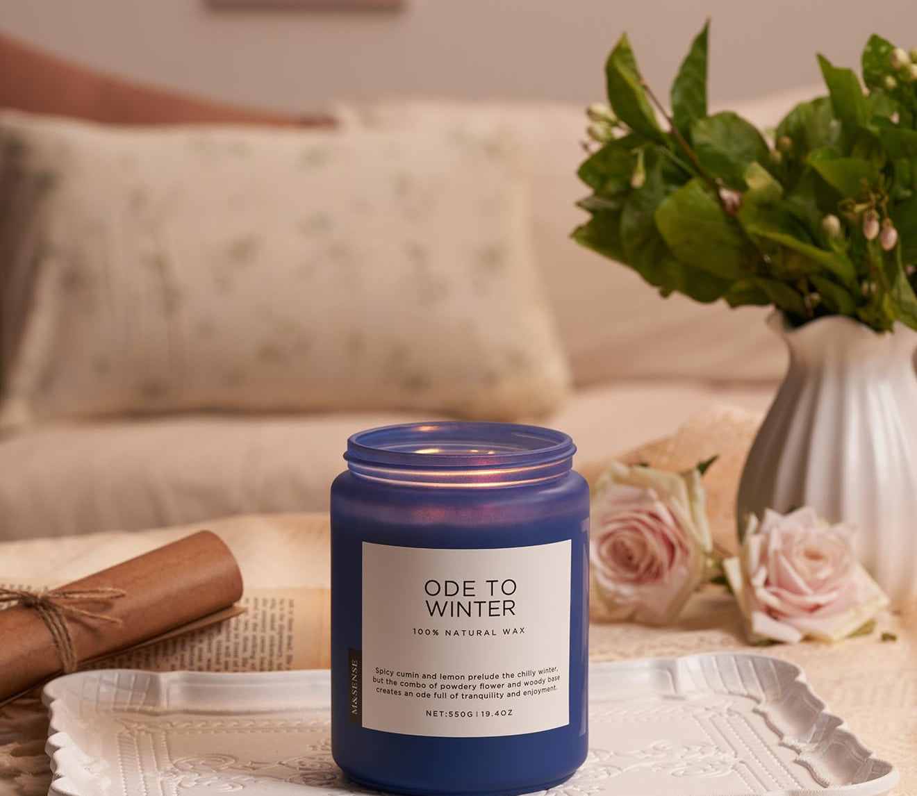 Blue candle labeled 'Ode to Winter' on a decorative tray with flowers and a blanket in the background.