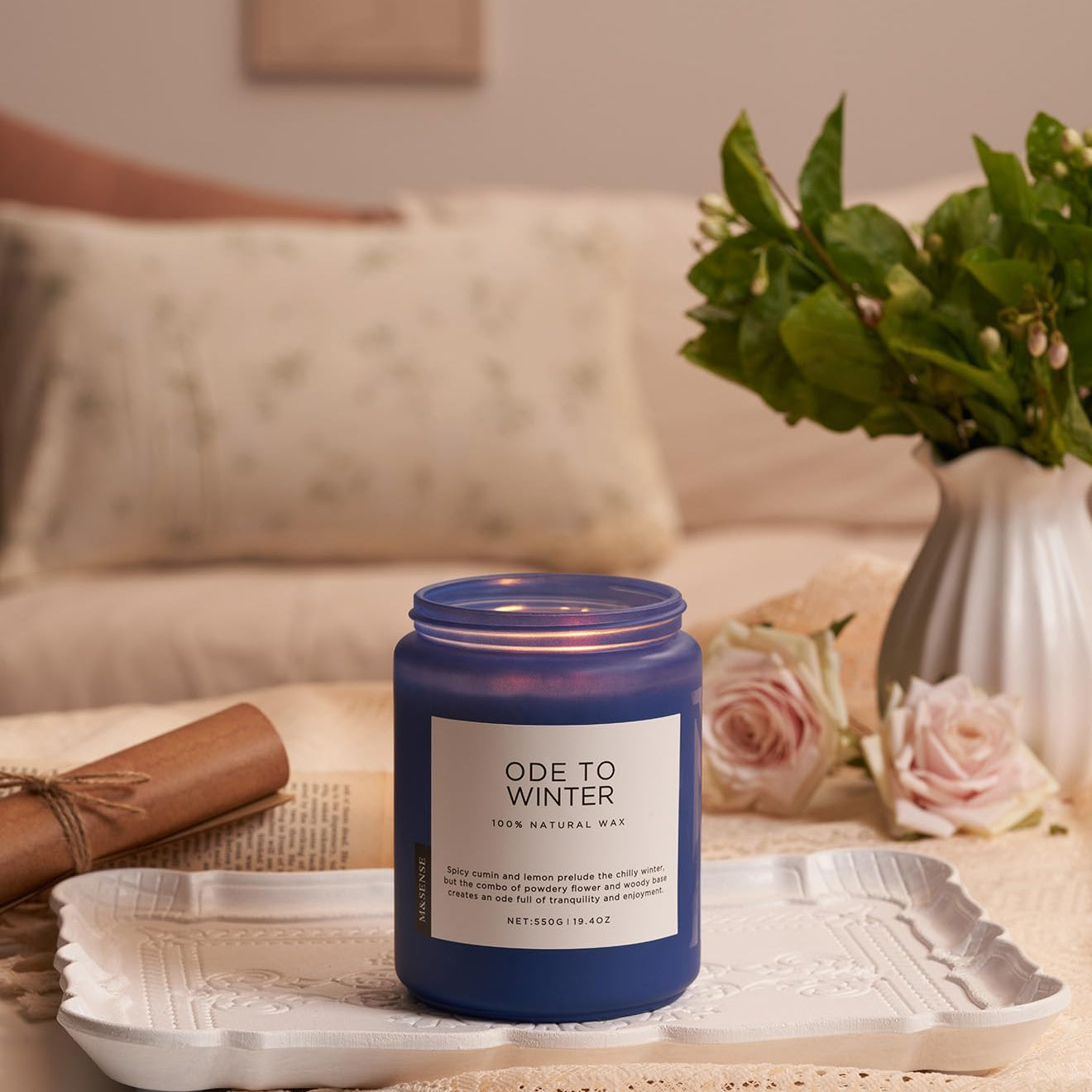 Blue candle labeled 'Ode to Winter' on a decorative tray with flowers and a blanket in the background.