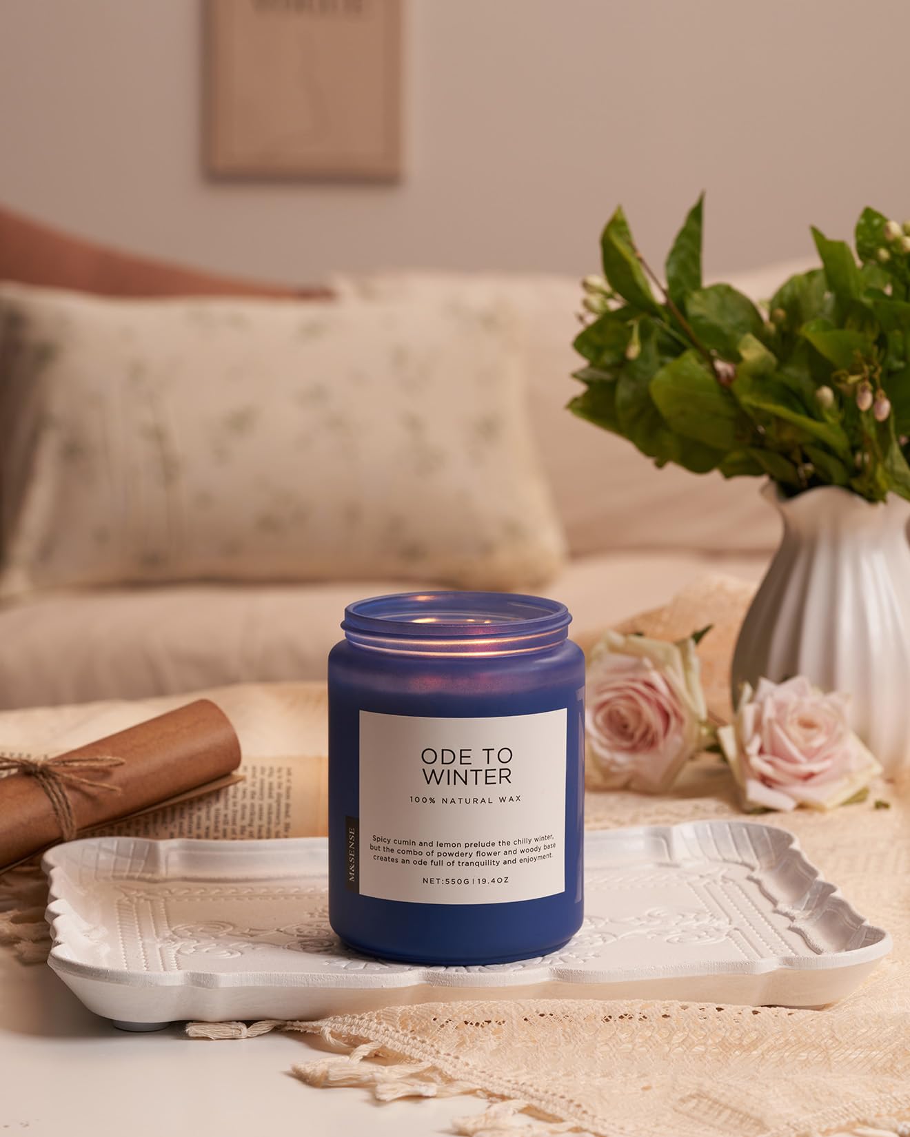 Blue candle labeled 'Ode to Winter' on a decorative tray with flowers and a blanket in the background.