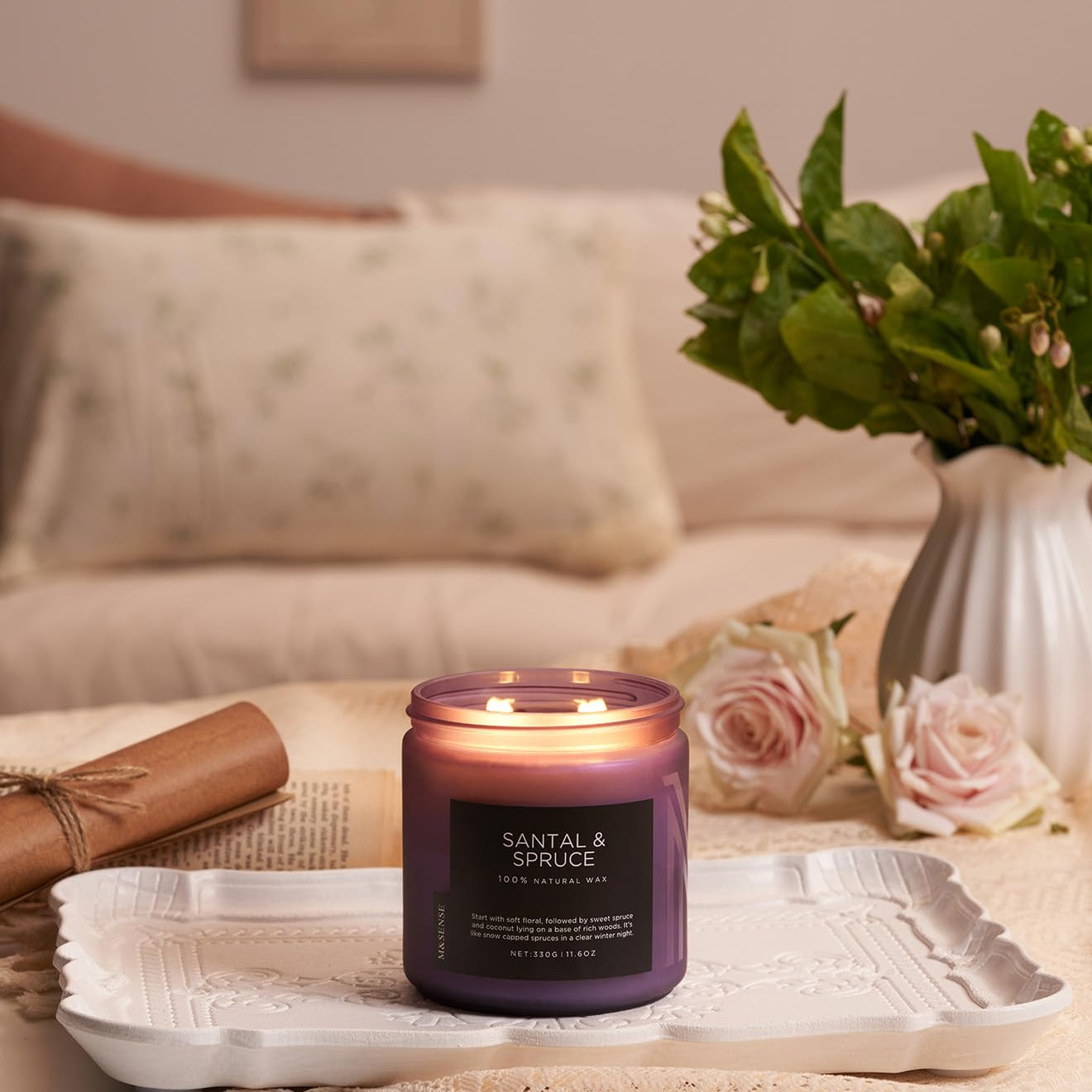 Scented candle labeled 'Santal & Spruce' on a tray with flowers and a vase in the background.
