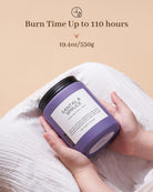 Hand holding a purple candle labeled 'Santal & Spruce' with text about burn time and weight on a beige background.
