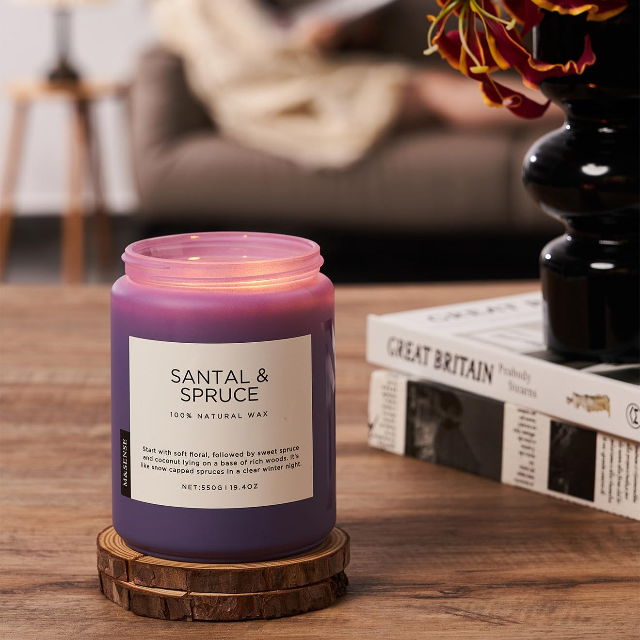 Purple candle labeled 'Santal & Spruce' on a wooden coaster with a blurred background of a person reading.