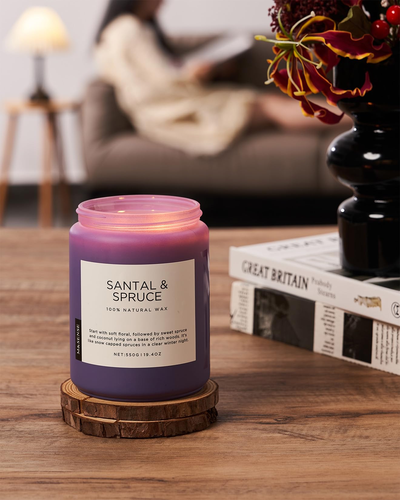Purple candle labeled 'Santal & Spruce' on a wooden coaster with a blurred background of a person reading.