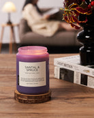 Purple candle labeled 'Santal & Spruce' on a wooden coaster with a blurred background of a person reading.