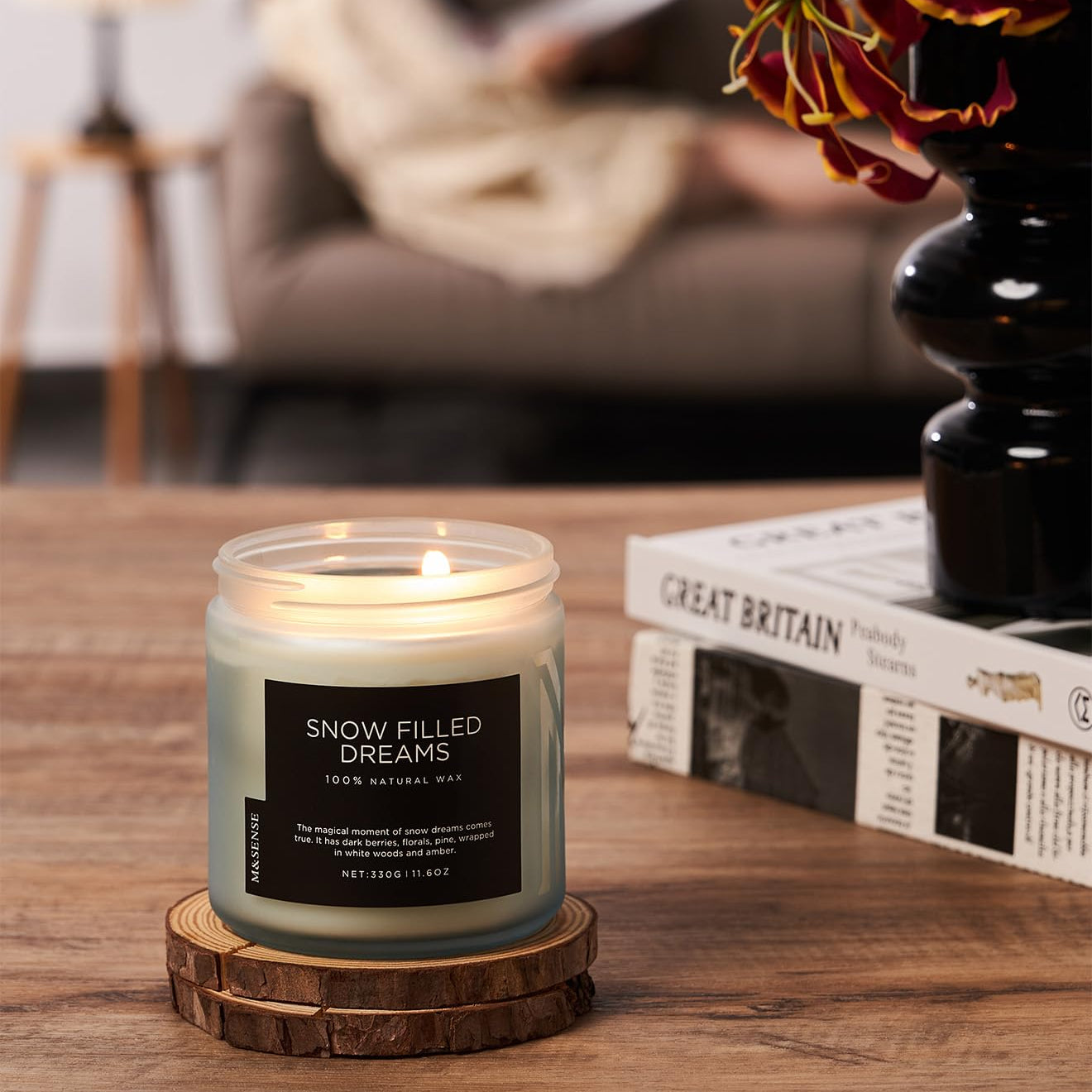 Candle labeled 'Snow Filled Dreams' on a wooden coaster with a blurred background of a person reading a book.