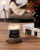 Candle labeled 'Snow Filled Dreams' on a wooden coaster with a blurred background of a person reading a book.
