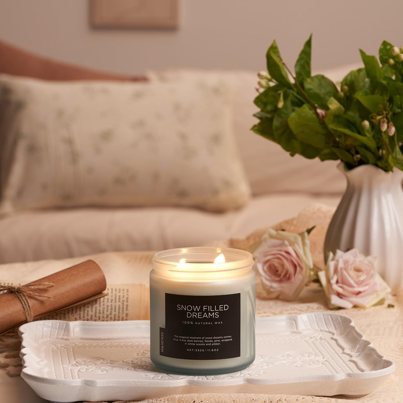 Candle labeled 'Snow Filled Dreams' on a table with a vase of flowers and a blanket in the background.