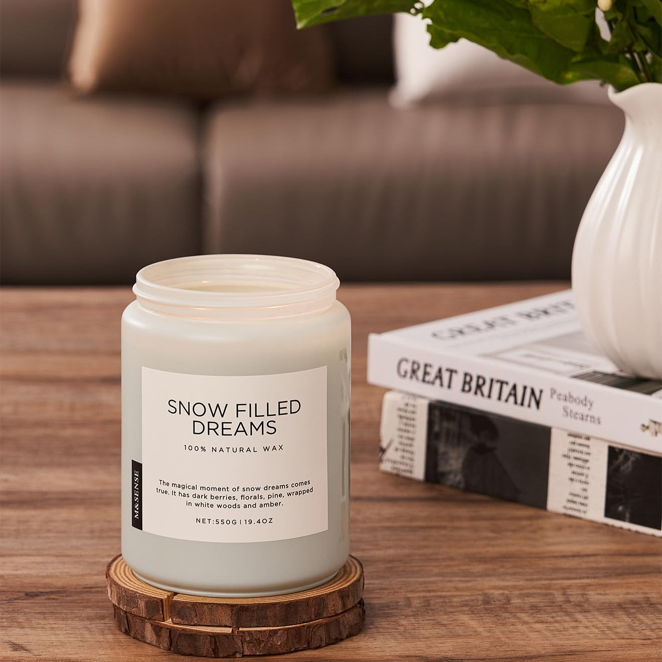 Candle labeled 'Snow Filled Dreams' on a wooden coaster with a vase and books in the background.