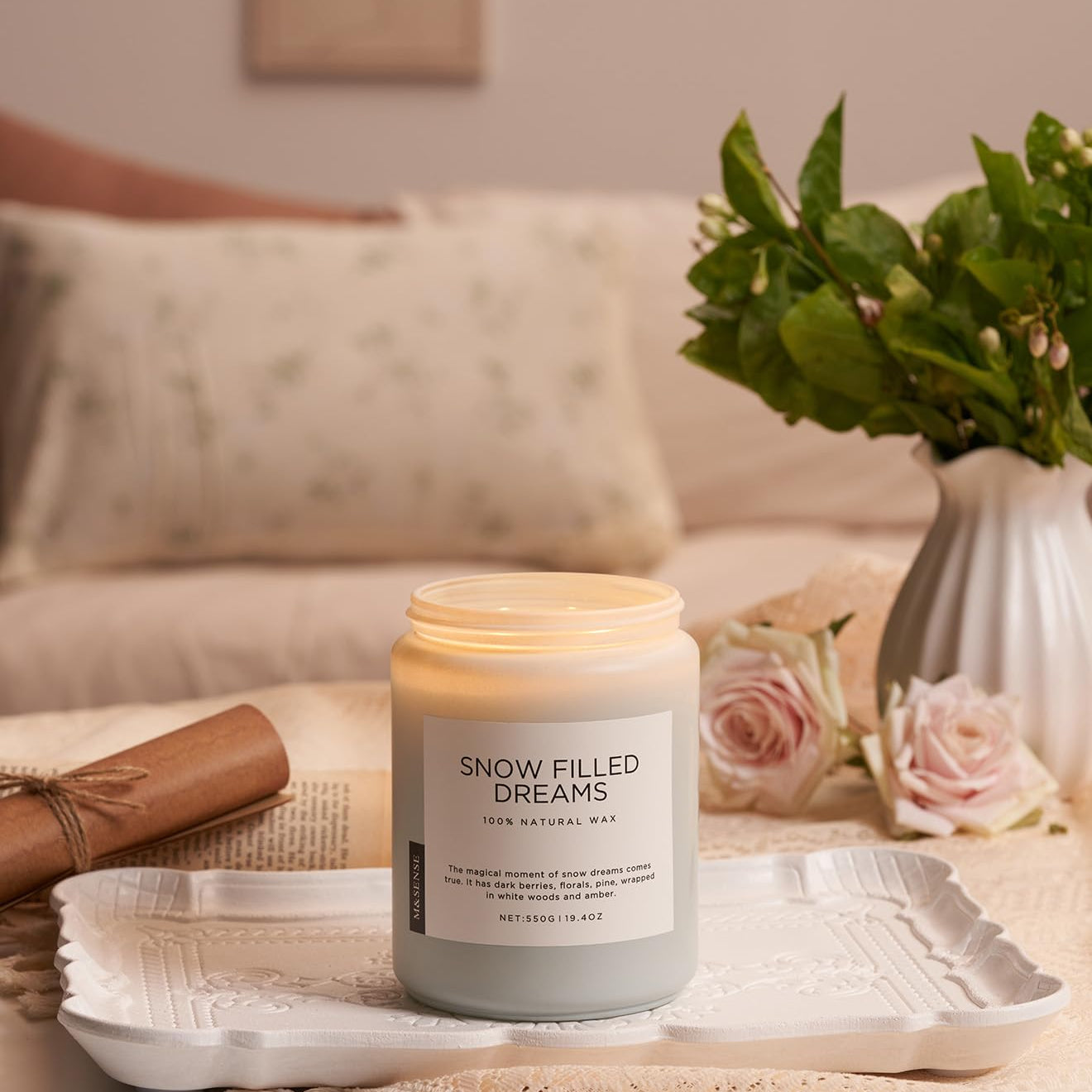 Candle labeled 'Snow Filled Dreams' on a table with a vase of flowers and a newspaper in the background.
