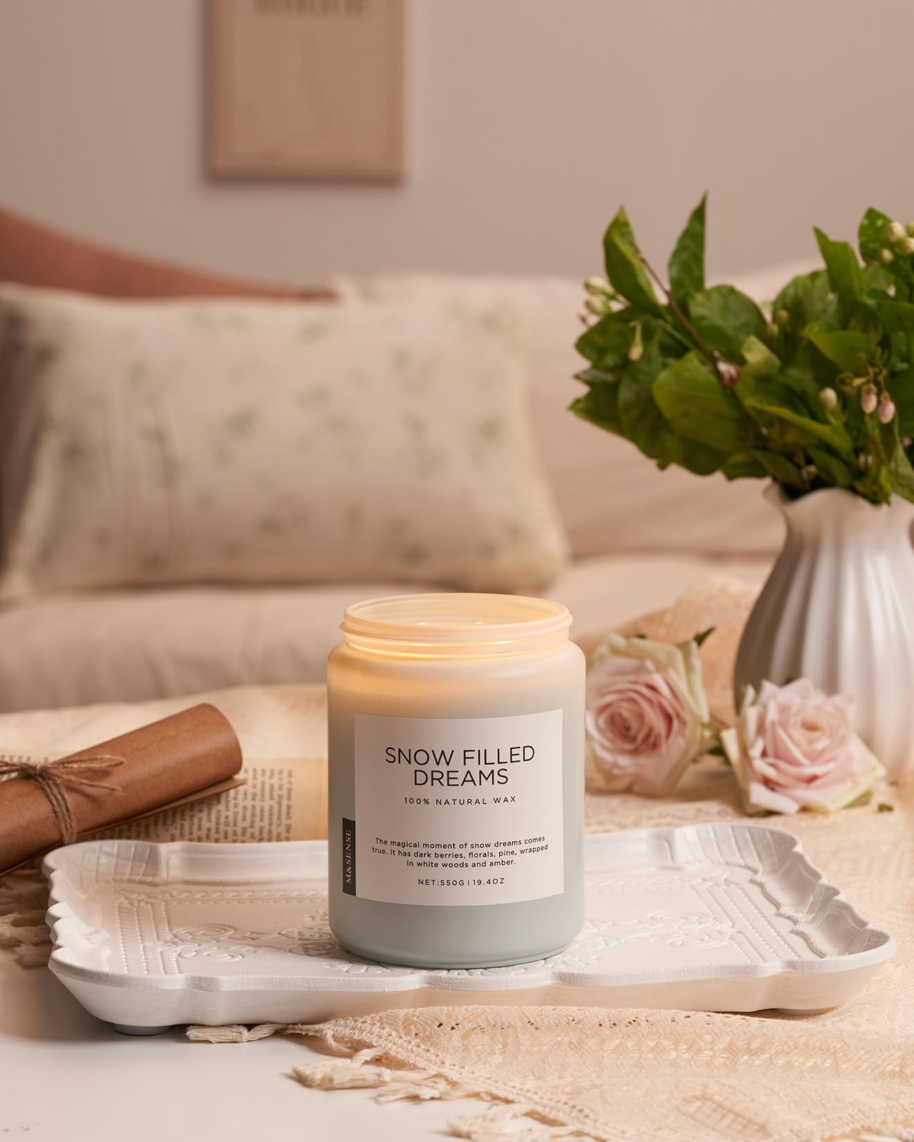 Candle labeled 'Snow Filled Dreams' on a table with a vase of flowers and a newspaper in the background.